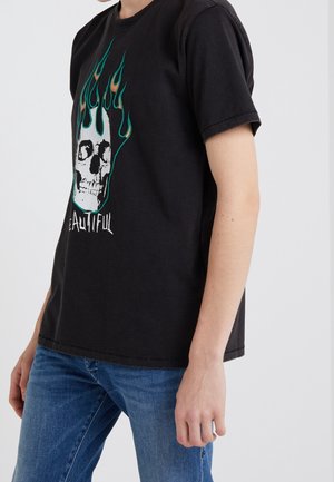Personne portant un T-shirt noir avec un crâne blanc entouré de flammes vertes et le mot "Beautiful" partiellement visible, associé à un jean bleu.