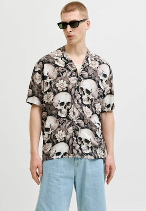 Młody mężczyzna noszący czarne okulary przeciwsłoneczne i koszulę na guziki z czarno-białym kwiatowym i czaszkowym wzorem, połączoną z jasnoniebieskimi dżinsami.