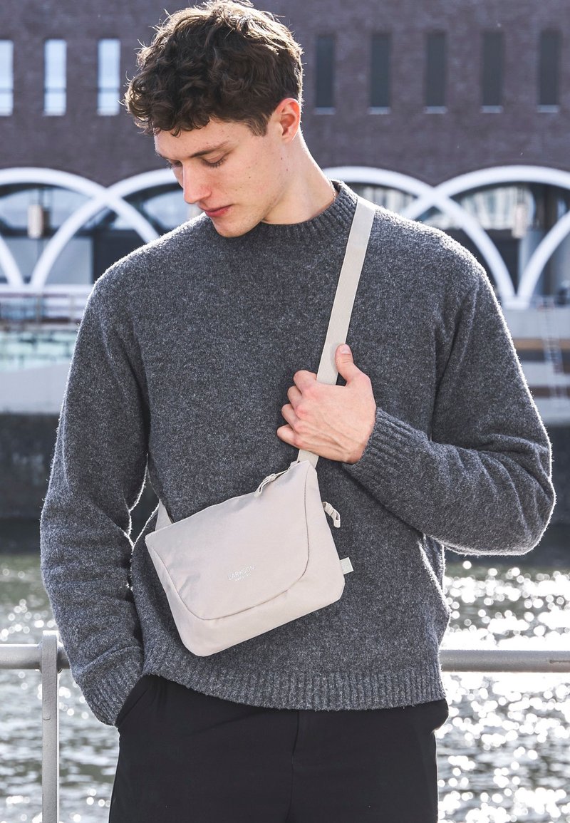 Světlé šedé crossbody taška s klopou a jemným logem značky; nošena přes tmavě šedý pletený svetr v řece.