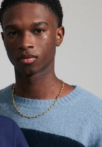 Hellblauer Wollpullover mit geripptem Halsausschnitt und einem dunkelblauen Streifen. Goldene Kettenhalskette mit ovalen Gliedern. Glatte Hautstruktur.