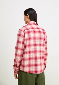 Rdeče in bele kvadratne flanelaste srajce z dolgimi rokavi, gumbnimi manšeti in ovratnikom. Nošene čez zelene hlače, ki imajo sproščen kroj in mehak texture.