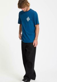 Hombre con cabello rubio, vestido con una camiseta gráfica azul, pantalones holgados negros y zapatillas negras, de pie frente a un fondo blanco.