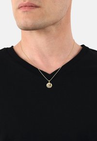 Gul hängande kedja på en silverkedja, med en cirkulär design med ett vågmönster och en graverad figur, bärs över en svart V-ringad skjorta.