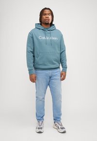 Moleton em algodão azul-petróleo com bolso frontal e capuz com cordão, apresentando o logo branco "Calvin Klein". Combinado com jeans azul claro e sapatilhas cinzentas.