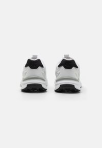 Bijele sportske tenisice s crnim naglaskom na peti, s teksturiranom potplatom, trakicom za povlačenje i logotipom na stražnjoj strani; aerodinamičan dizajn.