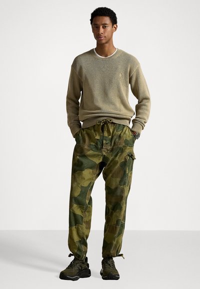 Beżowy sweter z dzianiny, zielone spodnie cargo w kamuflażu z elastycznym pasem i ciemne sneakersy. Teksturowany materiał i luźny krój.