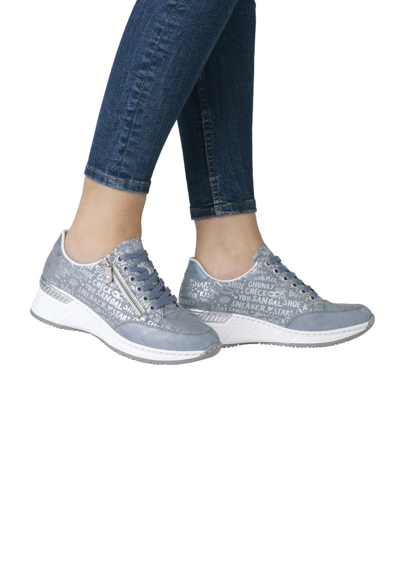 Gray suede sneakers with white text patterns, lace-up front, and a textured white rubber sole. Worn with dark blue fitted jeans.