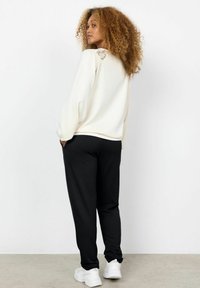Krämfärgad sweatshirt med spetsdetalj på baksidan, kombinerad med svarta joggers. Modellen bär vita sneakers och visar en avslappnad passform.