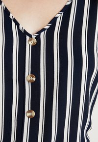 Tissu à rayures bleu marine et blanc avec un col en V ; comporte trois boutons ronds beige en accent sur le devant.