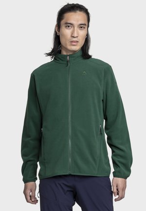 Mann mit langen dunklen Haaren, der eine dunkelgrüne Fleecejacke mit Reißverschluss und marineblaue Hose trägt, steht vor einem schlichten hellen Hintergrund.