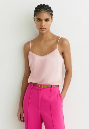 Punaseppe punutud juustega naine kannab heleroosa maika, mis on sarkaga sisse toppitud, ning helekollaste kõrge vöökohaga püksidega, millel on õhuke pruun vöö, seistes valge seina taustal.