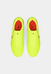 Zapatillas de fútbol en un brillante color amarillo con superficie texturizada y acentos rojos, con cordones blancos y un logo negro de Nike en el costado.