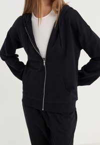 Sweat à capuche noir zippé en tissu doux, avec une capuche, des poches avant et des poignets côtelés. Porté sur un haut blanc côtelé.