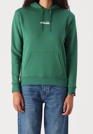 Person trägt grünen ellesse-Kapuzenpullover mit Fronttasche und blaue Jeans, steht vor hellem einfarbigem Hintergrund.