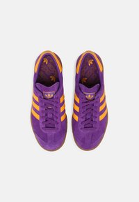 Purple suede athletic shoes with orange stripes, featuring a lace-up design and a textured gum rubber sole. Interior displays Adidas branding.