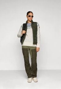 Olivgrüne Steppweste über einem gemusterten weißen Pullover, kombiniert mit olivgrünen Cargo-Hosen mit Taschen und hellen Schuhen. Als Accessoires Sonnenbrille.