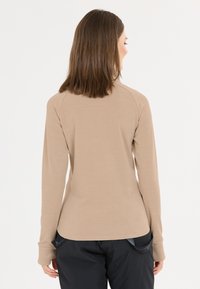 Kvinde med skulderlangt brunt hår iført en beige langærmet bluse og sorte bukser, set fra bagenden mod en hvid baggrund.