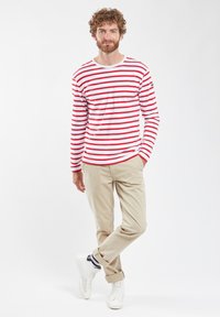 Camicia a maniche lunghe a righe con righe rosse e bianche, in tessuto di cotone, con scollatura rotonda. Abbinata a pantaloni beige e sneakers bianche.