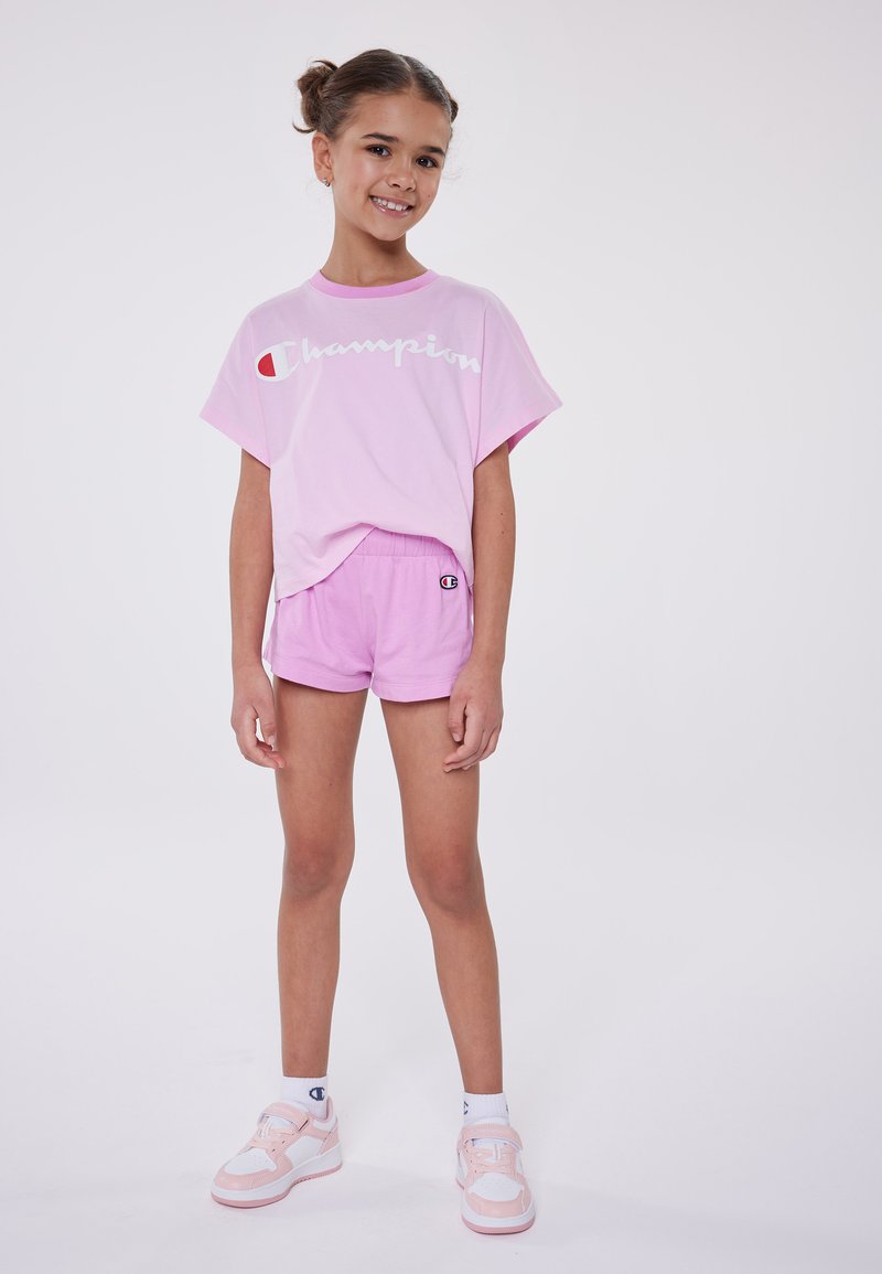Camiseta de algodón rosa con el logo blanco de "Champion", combinada con unos pantalones cortos rosa a juego. Zapatillas deportivas blancas y rosas completan el conjunto.