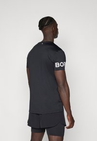 Schwarzes sportliches T-Shirt mit rundem Hals und kurzen Ärmeln, mit dem Wort "BORN" in Weiß auf dem linken Ärmel gedruckt. Schlanke Passform.