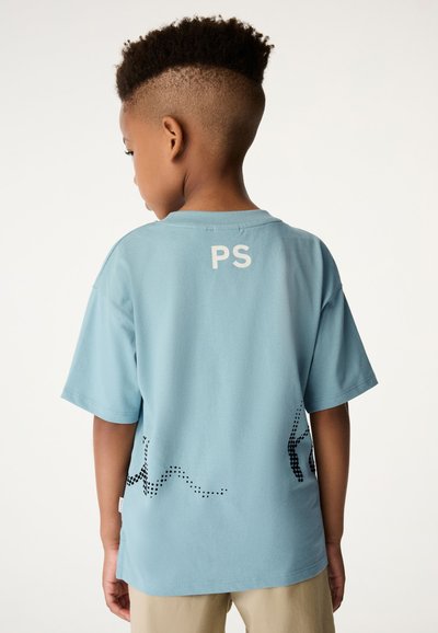 Gaiši zils kokvilnas T-krekls ar īsām piedurknēm, ar baltiem "PS" uzrakstiem aizmugurē un melnu punktiņu rindas dizainu tuvu apakšējai malai.