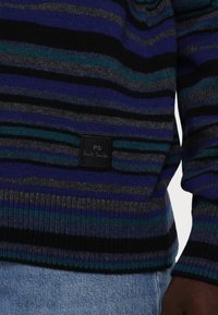 Gros plan d'une personne portant un pull à rayures bleues, vertes, noires et grises avec une étiquette noire PS Paul Smith au-dessus d'un jean bleu.