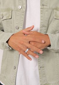Anello d'argento indossato sulle dita, con un design liscio e allungato. Le mani sono unite, indossando una giacca verde chiaro sopra una camicia bianca.