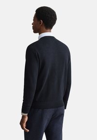 Dunkelblauer Pullover mit Rundhalsausschnitt, gerippten Bündchen und Saum. Wird über einem weißen Hemd getragen, zeichnet sich durch glatte Textur und minimalistisches Design aus.