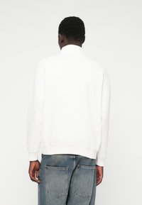 Baltas kreklu ar apaļu apkakli, brīvi pieguļošs, ar ribām pie piedurknēm un apakšmalas. Valkāts ar gaiši ziliem džinsiem, kuriem ir divas aizmugures kabatas.