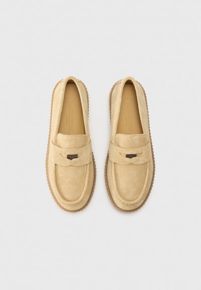 Mocassins en daim beige avec un bout arrondi, présentant une sangle décorative et un détail de logo. La semelle est texturée et en caoutchouc.
