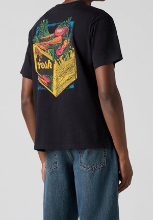 T-shirt noir avec un graphisme coloré d'une caisse en bois remplie de carottes fraîches, de tomates et de verdure dans le dos, porté avec un jean bleu.