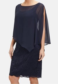 Femme portant une robe jupe en dentelle bleu marine avec un top à superposition transparente, manches fendues ouvertes et ourlet asymétrique.