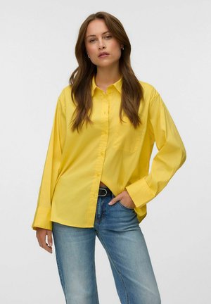Femme debout, une main dans la poche, portant une chemise jaune ample boutonnée et un jean bleu clair sur un fond clair uni.