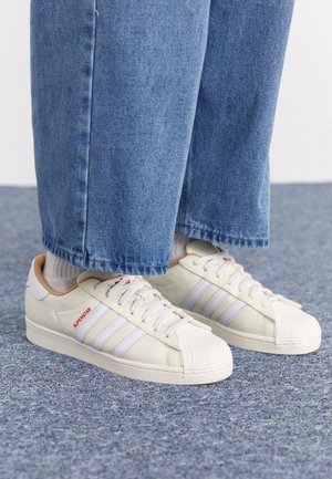 Stopy w białych butach Adidas Superstar z czerwonym napisem, połączone z białymi skarpetkami i niebieskimi dżinsowymi spodniami na szarym wykładzinowym podłożu.