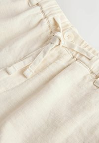Nahaufnahme des Bunds von cremefarbenen Baumwollhosen mit elastischem Bund, Knopf, verstellbarem Stoffbindeband und Details der Fronttasche.