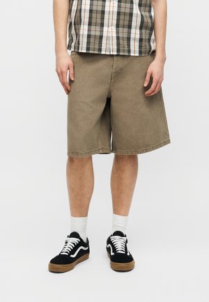 Bărbat purtând pantaloni scurți bej până la genunchi, o cămașă în carouri cu mâneci scurte, șosete albe și adidași negri cu șireturi albe, pe un fundal simplu.