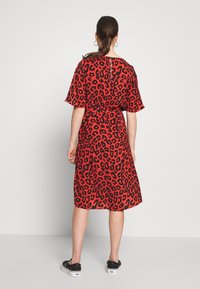 Robe rouge avec imprimé léopard noir, manches courtes et taille cintrée. Présente un détail de dos en forme de clé. Portée avec des baskets noires.