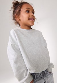 Világosszürke pulóver kerek nyakkal, laza fazonban, felhajtott ujjakkal, magasderekú szürke rövidnadrággal párosítva, semleges háttér előtt.