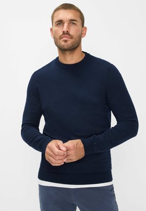 Marineblauwe gebreide trui met lange mouwen, ronde hals en geribbelde manchetten. Glad textuur, klassieke pasvorm, gelaagd over een wit shirt.
