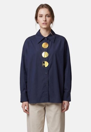 Jeune femme portant une chemise oversize bleu foncé avec trois grands boutons ronds dorés brillants et un pantalon beige, debout devant un fond uni.