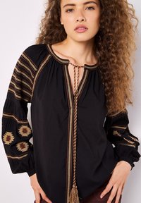 Blusa nera con maniche lunghe, ricami decorativi in oro e marrone, scollatura a chiave e un laccio intrecciato con dettaglio a frangia.