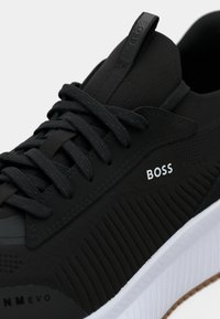 Sneaker BOSS nera con tessuto testurizzato, lacci neri e suola bianca in primo piano che mostra il logo sul lato e il testo "NM EVO" vicino alla punta.