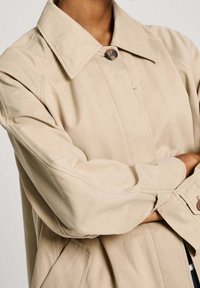 Beige sako se strukturovaným límcem, zapínáním na jeden knoflík a mírně uvolněným střihem. Má hladkou texturu a jemné detaily prošití.