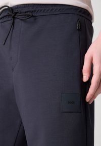 Marinblå joggerbyxor tillverkade av mjukt tyg, med dragsko i midjan, sidofickor och en fyrkantig logotyp på vänster ben.