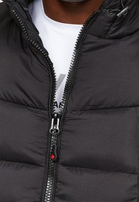 Schwarze gesteppte Weste mit hohem Kragen, Reißverschluss mit silbernem Zipper und rotem Logo-Akzent. Texturiertes Material mit horizontalen Steppnähten.