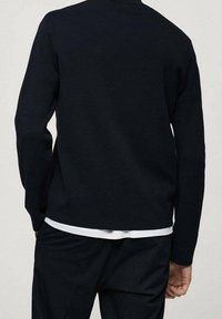 Pull en maille bleu marine avec un col rond, des manches longues et une texture lisse, porté sur un sous-vêtement blanc, accompagné d'un pantalon noir.