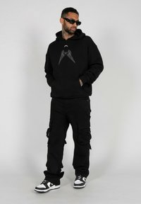 Zwarte hoodie met grafisch ontwerp, zwarte cargobroek en zwart-witte sneakers. Accessoires omvatten een zonnebril. Over het geheel een gladde textuur.