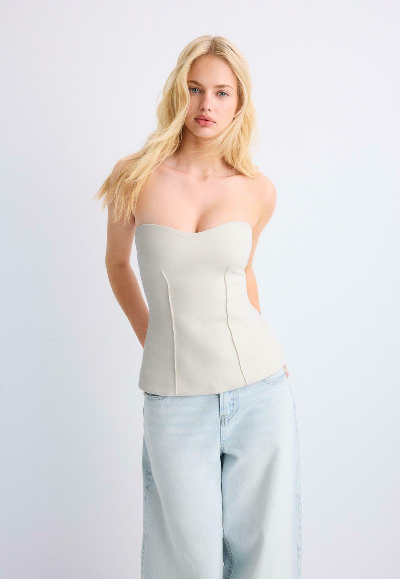 Jeune femme aux longs cheveux blonds portant un haut beige sans bretelles et un jean large bleu clair, sur un fond uni.