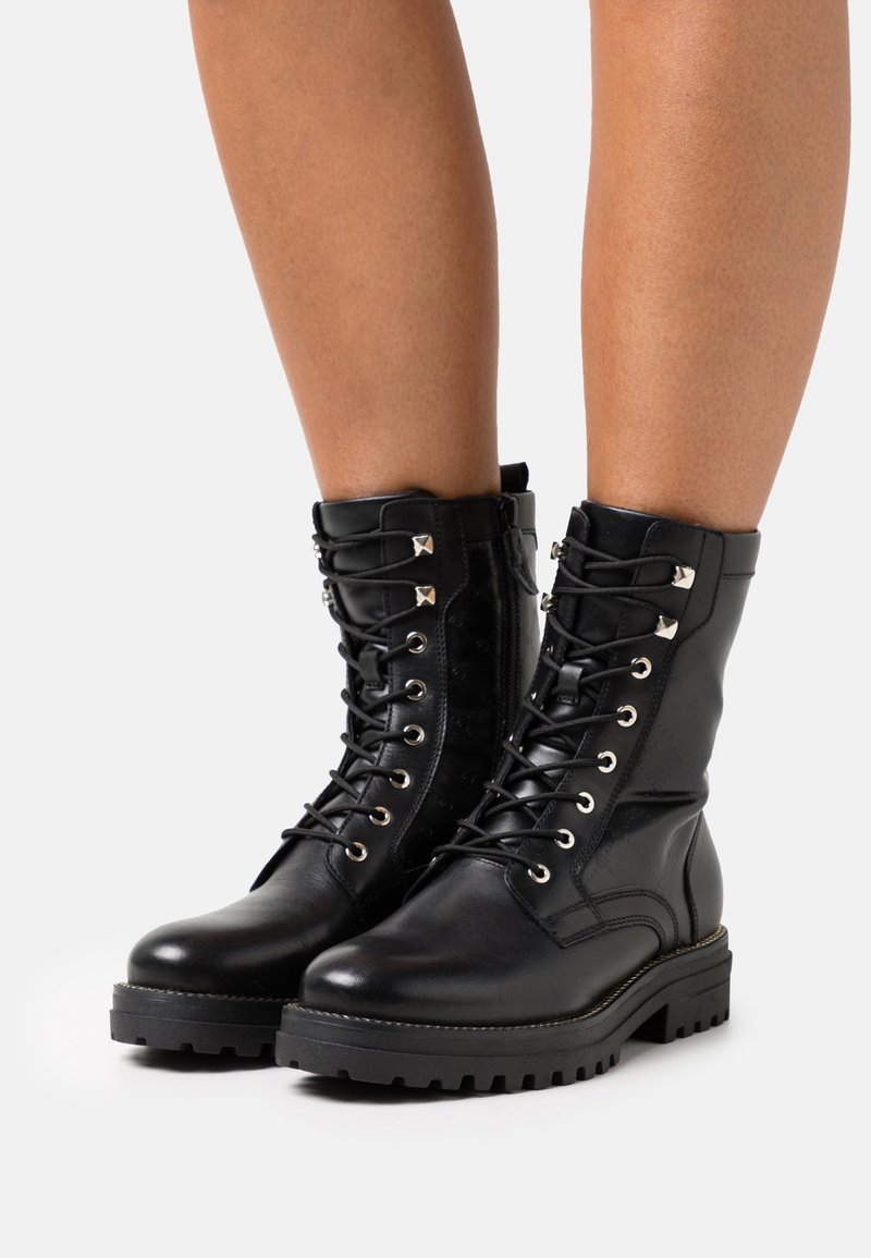 Bottes de combat noires en cuir à lacets avec œillets métalliques et clous portées sur des jambes nues sur un fond blanc uni.