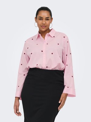 Sieviete, kas valkā rozā kreklu ar sarkaniem sirsniņu rakstiem un melnu zīmuļsvārku, stāvot pret vienkāršu gaišu fonu.